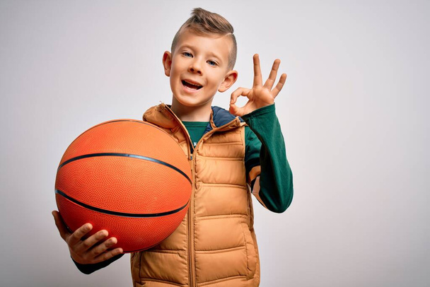 Young little caucasian sports kid playing basketball holding orange ball over isolated background doing ok sign with fingers, excellent symbol - Photo, Image