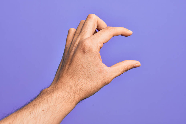Hand van Kaukasische jongeman met vingers over geïsoleerde paarse achtergrond plukken en nemen onzichtbare ding, houden object met vingers tonen ruimte - Foto, afbeelding