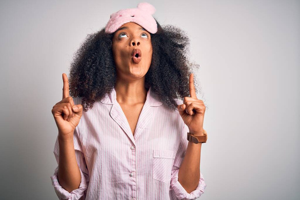 Joven mujer afroamericana con cabello afro usando máscara para dormir y pijama rosa asombrada y sorprendida mirando hacia arriba y señalando con los dedos y los brazos levantados
. - Foto, Imagen