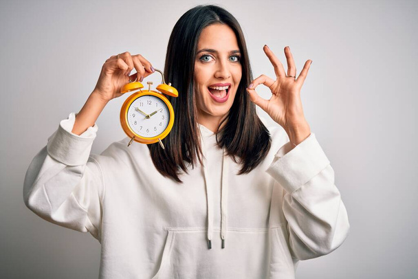 Young woman with blue eyes holding alarm clock standing over isolated white background doing ok sign with fingers, excellent symbol - Фото, зображення