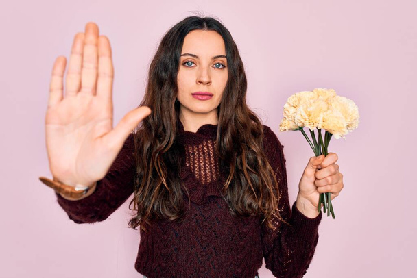 Young beautiful woman with blue eyes holding bouquet of yellow flowers over pink background with open hand doing stop sign with serious and confident expression, defense gesture - Foto, Imagen
