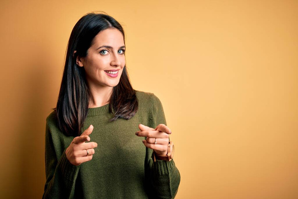 Young brunette woman with blue eyes wearing green casual sweater over yellow background pointing fingers to camera with happy and funny face. Good energy and vibes. - Foto, Bild