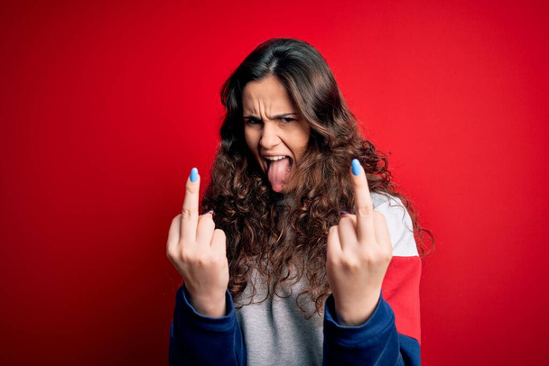 Young beautiful woman with curly hair wearing casual sweatshirt over isolated red background Showing middle finger doing fuck you bad expression, provocation and rude attitude. Screaming excited - Foto, Bild
