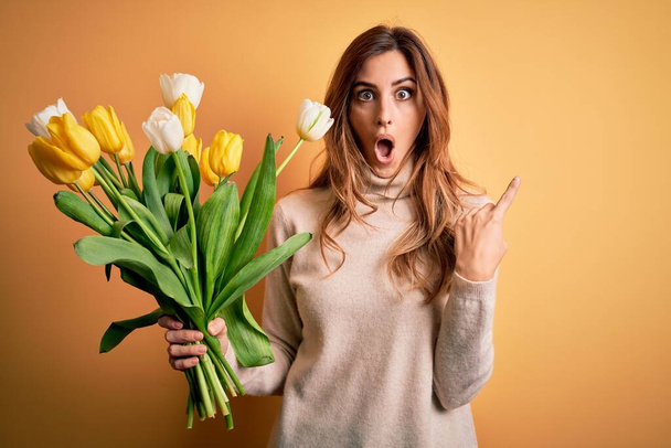 Młoda piękna brunetka trzyma bukiet żółtych tulipanów na odizolowanym tle Zaskoczony wskazując palcem w bok, otwarte usta zdumiony wyraz. - Zdjęcie, obraz