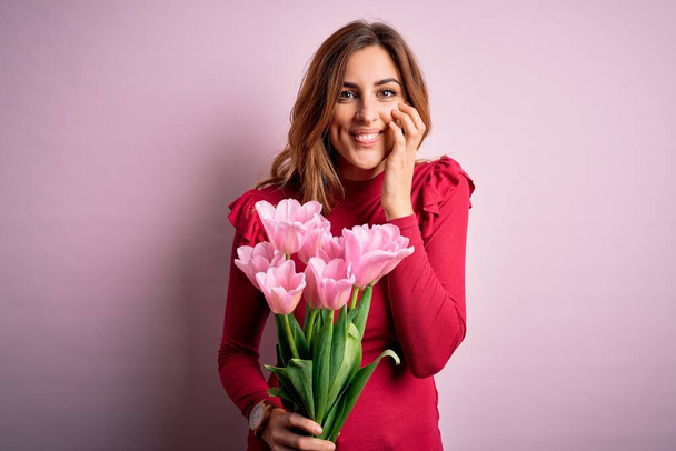 Junge schöne brünette Frau mit einem Strauß rosa Tulpen vor isoliertem Hintergrund lacht und verlegen kichert, den Mund mit Händen bedeckt, Klatsch und Skandalkonzept - Foto, Bild