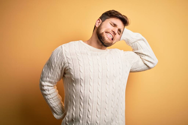 Young blond man with beard and blue eyes wearing white sweater over yellow background stretching back, tired and relaxed, sleepy and yawning for early morning - Photo, Image