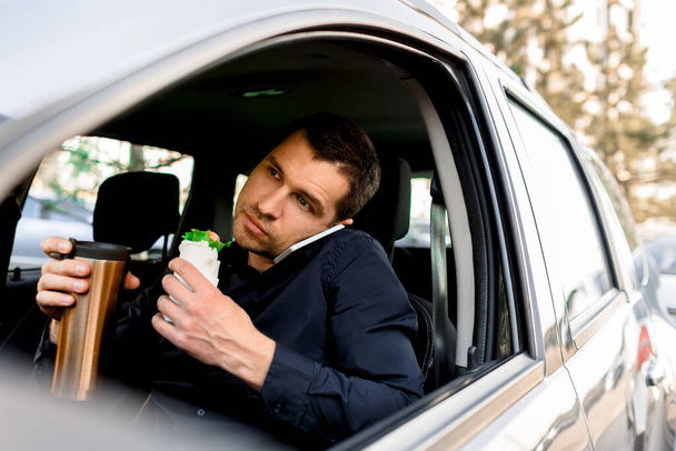 Зупинка укусу, щоб поїсти. Чоловік їсть закуску в машині і п'є каву або чай
. - Фото, зображення