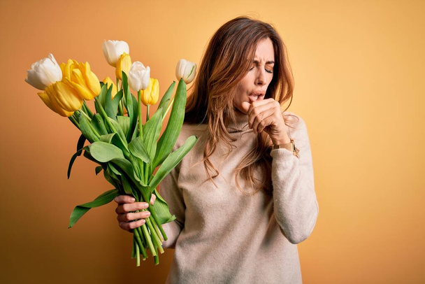 Young beautiful brunette woman holding bouquet of yellow tulips over isolated background feeling unwell and coughing as symptom for cold or bronchitis. Health care concept. - Photo, Image