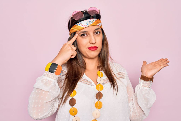 Jonge Spaanse hippie vrouw dragen mode boho stijl en een zonnebril over roze achtergrond verward en geïrriteerd met open palm tonen kopieerruimte en wijzende vinger naar het voorhoofd. Denk er eens over na.. - Foto, afbeelding