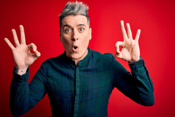 Young handsome modern man wearing elegant green shirt over red isolated background looking surprised and shocked doing ok approval symbol with fingers. Crazy expression - Photo, Image