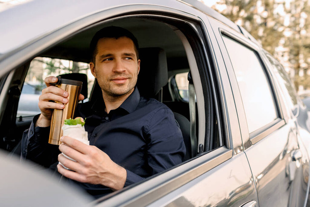 Parando a comer algo. El hombre come bocadillos en el coche y bebe café o té
. - Foto, Imagen