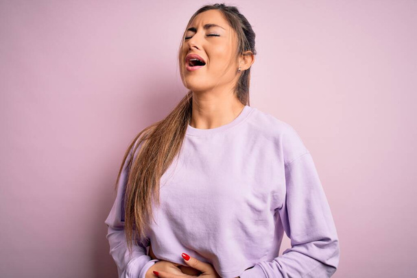 吐き気、痛みを伴う病気が具合が悪いので、若い美しいスポーツ女性は胃の上に手でピンクの隔離された背景にスウェットシャツを着て。アチェの概念. - 写真・画像