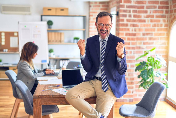 Middle age handsome businessman wearing glasses sitting on desk at the office celebrating surprised and amazed for success with arms raised and open eyes. Winner concept. - Фото, зображення