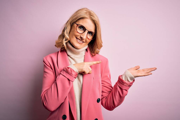 Красивая блондинка среднего возраста, одетая в элегантный розовый пиджак и очки, поражена и улыбается в камеру, представляя рукой и указывая пальцем
. - Фото, изображение