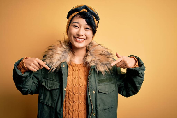 Beautiful asian skier girl wearing snow sportswear using ski goggles over yellow background looking confident with smile on face, pointing oneself with fingers proud and happy. - Photo, Image
