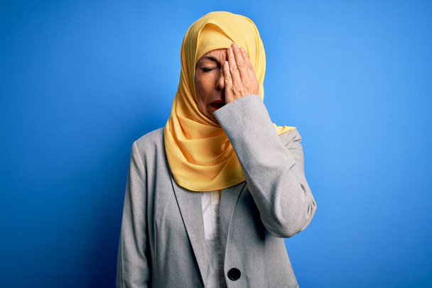 Μεσήλικας μελαχρινή επιχειρηματίας γυναίκα φορώντας muslim παραδοσιακή μαντίλα πάνω από μπλε φόντο Χασμουρητό κουρασμένος καλύπτει το μισό πρόσωπο, το μάτι και το στόμα με το χέρι. Το πρόσωπο πονάει από τον πόνο. - Φωτογραφία, εικόνα
