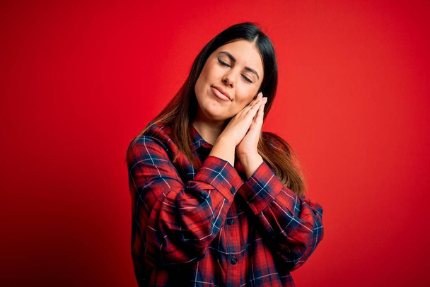 Young beautiful woman wearing casual shirt over red background sleeping tired dreaming and posing with hands together while smiling with closed eyes. - 写真・画像