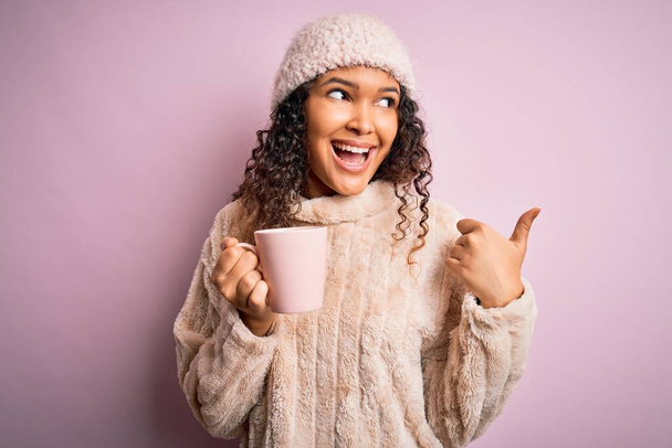 Jonge mooie vrouw met krullend haar drinken mok koffie over geïsoleerde roze achtergrond wijzen en tonen met duim naar de zijkant met gelukkig gezicht glimlachen - Foto, afbeelding