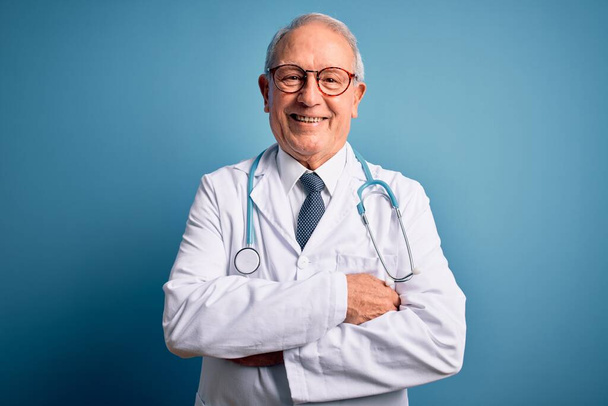 Starszy siwowłosy lekarz noszący stetoskop i płaszcz medyczny na niebieskim tle szczęśliwa twarz uśmiechnięta skrzyżowanymi ramionami patrzącymi w kamerę. Osoba pozytywna. - Zdjęcie, obraz