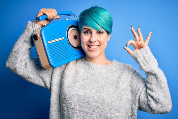 Jonge vrouw met blauwe mode haar luisteren naar muziek met vintage draagbare radio doet ok teken met de vingers, uitstekend symbool - Foto, afbeelding