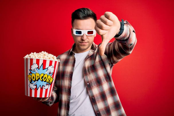 Nuori komea valkoihoinen mies yllään 3D-elokuvalasit ja syö popcornia etsivät onneton ja vihainen osoittaa hylkääminen ja negatiivinen peukalot alas ele. Huono ilme
. - Valokuva, kuva