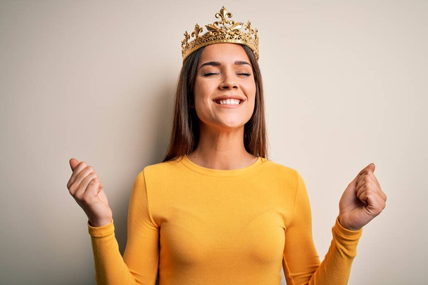 Young beautiful brunette woman wearing golden queen crown over white background very happy and excited doing winner gesture with arms raised, smiling and screaming for success. Celebration concept. - Photo, Image