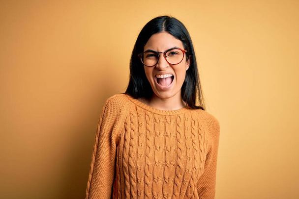 Jeune belle femme hispanique portant des lunettes sur fond jaune isolé clin d'oeil en regardant la caméra avec une expression sexy, gai et heureux visage
. - Photo, image