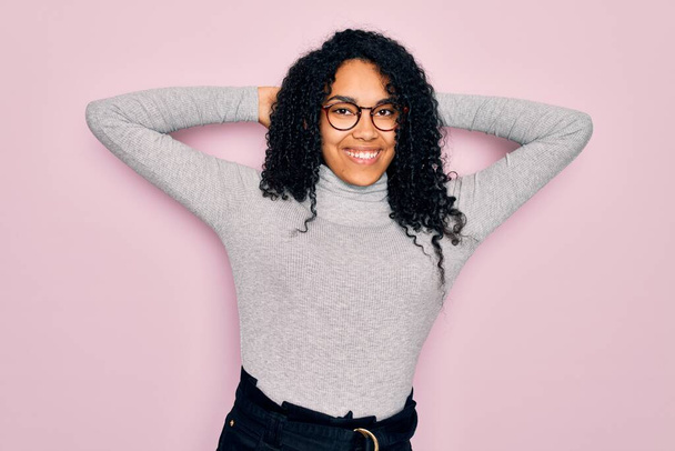 Jonge Afro-Amerikaanse vrouw draagt coltrui en bril over roze achtergrond ontspannen en stretching, armen en handen achter hoofd en nek glimlachend gelukkig - Foto, afbeelding