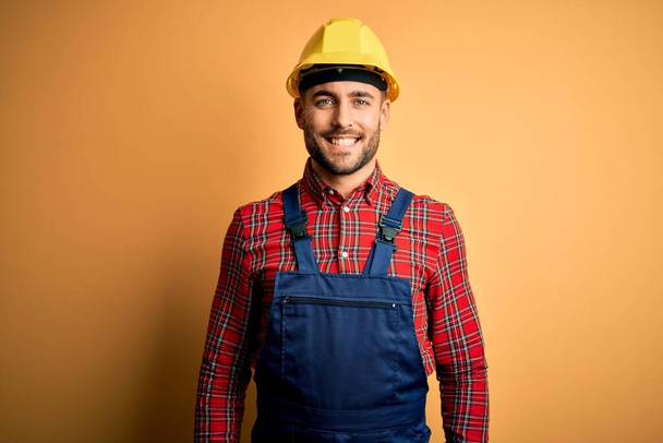 Молодой строитель в строительной форме и шлеме безопасности на желтом изолированном фоне со счастливой и прохладной улыбкой на лице. Счастливчик
. - Фото, изображение