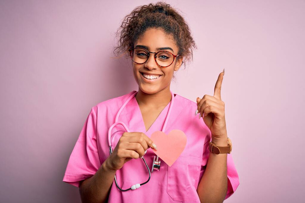 Chica cardióloga afroamericana con uniforme médico y estetoscopio sosteniendo el corazón sorprendido con una idea o pregunta apuntando con el dedo con la cara feliz, número uno
 - Foto, imagen