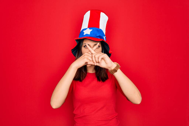 Fiatal gyönyörű barna nő visel egyesült államokbeli kalap ünnepli függetlenség napja Elutasítás kifejezés keresztbe ujjak csinál negatív jel - Fotó, kép