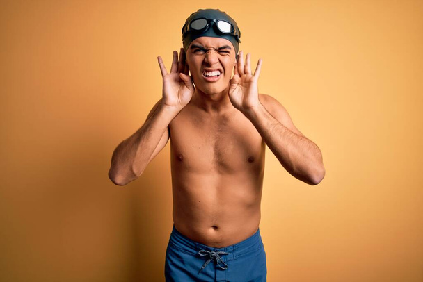 Молодой красивый мужчина без рубашки в купальнике и плавательной шапочке на изолированном желтом фоне пытается услышать жесты обеих рук, любопытные для сплетен. Проблемы со слухом, глухой
 - Фото, изображение