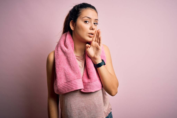 Młoda piękna brunetka sportowa ubrana w odzież sportową i ręcznik na różowym tle ręka na ustach opowiadając tajne plotki, szepcząc złośliwe rozmowy - Zdjęcie, obraz