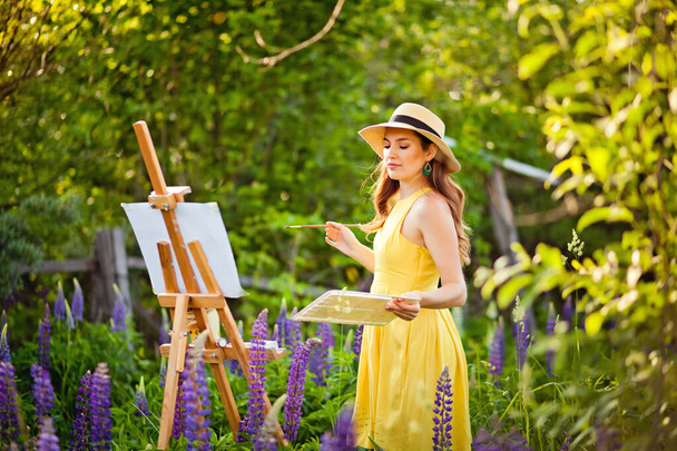 Una artista con un vestido amarillo y sombrero dibuja un campo de altramuz al aire libre. Caja de caballete con lienzo, pintura al óleo, paleta y herramientas de artista
. - Foto, Imagen