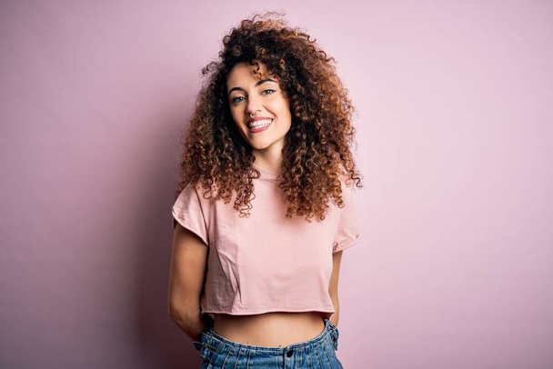 Young beautiful woman with curly hair and piercing wearing casual pink t-shirt with a happy and cool smile on face. Lucky person. - Foto, afbeelding