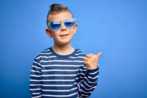 Νεαρό μικρό καυκάσιο παιδί με μπλε μάτια στέκεται φορώντας γυαλιά ηλίου πάνω από το μπλε φόντο χαμογελώντας με χαρούμενο πρόσωπο κοιτάζοντας και δείχνοντας προς τα πλάγια με τον αντίχειρα προς τα πάνω. - Φωτογραφία, εικόνα