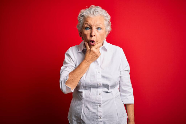 Ältere schöne Frau in elegantem Hemd steht über isoliertem rotem Hintergrund und schaut fasziniert mit Ungläubigkeit, Überraschung und erstauntem Ausdruck mit den Händen am Kinn - Foto, Bild
