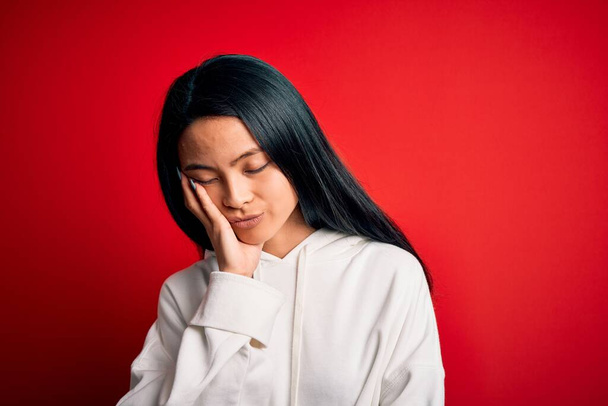 Jeune belle femme sportive chinoise portant un sweat-shirt sur fond rouge isolé pensant fatigué et ennuyé par des problèmes de dépression avec les bras croisés
. - Photo, image