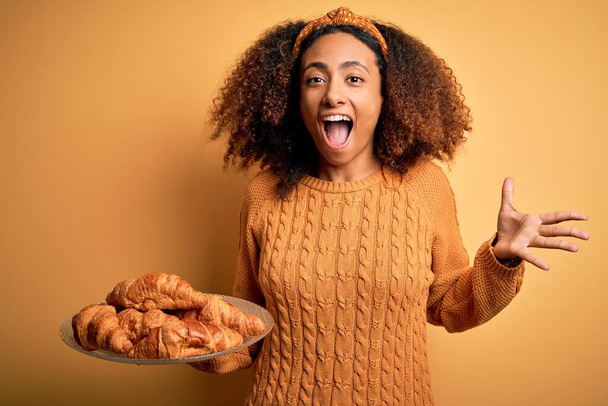 Junge afrikanisch-amerikanische Frau mit Afro-Haaren, die Croissants über gelbem Hintergrund hält, sehr glücklich und aufgeregt, Siegerausdruck, der den Sieg schreiend mit großem Lächeln und erhobenen Händen feiert - Foto, Bild