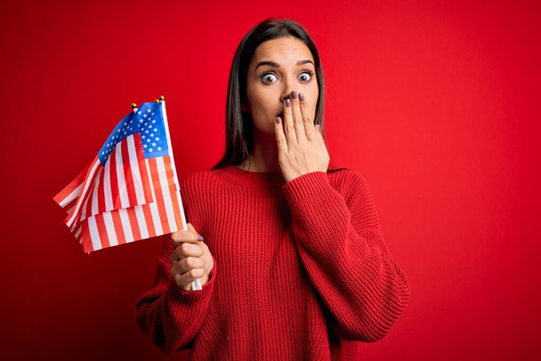 Νεαρή όμορφη μελαχρινή πατριωτική γυναίκα που κρατά την σημαία των Ηνωμένων Πολιτειών πάνω από το κόκκινο φόντο καλύπτουν το στόμα με το χέρι συγκλονισμένος από ντροπή για λάθος, έκφραση του φόβου, φοβάται στη σιωπή, μυστική έννοια - Φωτογραφία, εικόνα