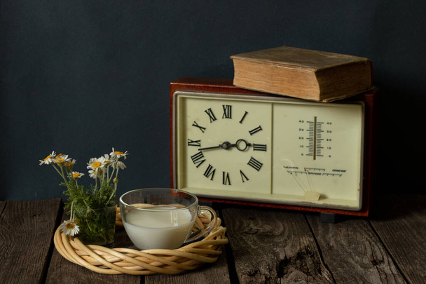 taza de leche y un jarrón con margaritas cerca del reloj con un libro sobre una mesa de madera
 - Foto, imagen