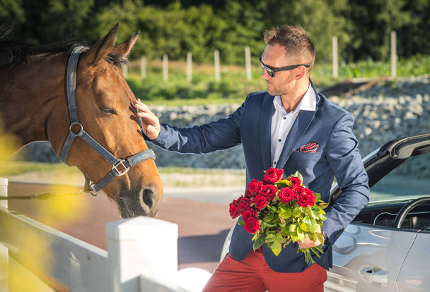 Gazdag jól öltözött fehér férfi kezében csokor vörös rózsa és simogató ló.  - Fotó, kép