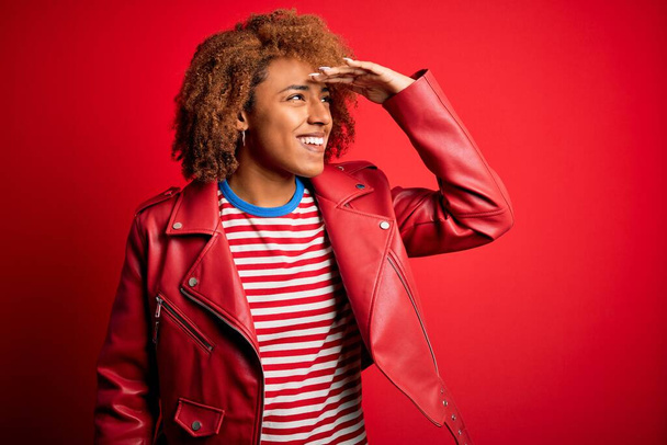 Young beautiful African American afro woman with curly hair wearing casual red jacket very happy and smiling looking far away with hand over head. Searching concept. - Фото, зображення