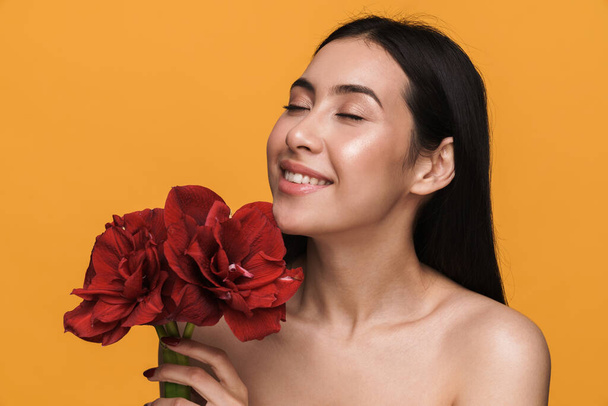 Beauty portrait of caucasian young brunette half-naked woman smiling and holding flowers isolated over yellow background - Foto, afbeelding