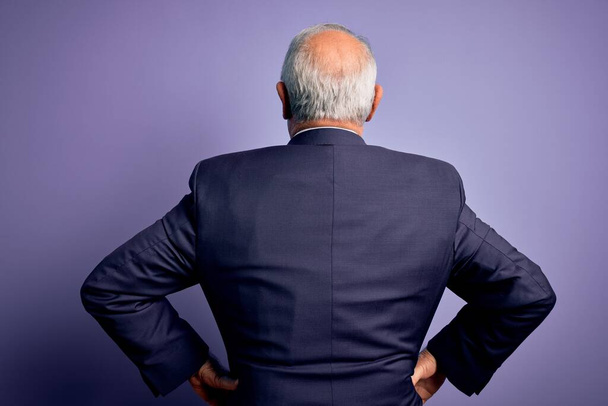 Grey haired senior business man wearing glasses and elegant suit and tie over purple background standing backwards looking away with arms on body - Fotografie, Obrázek