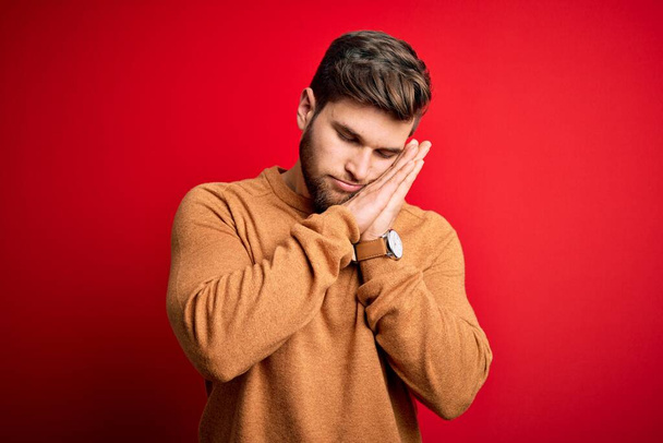 Young blond man with beard and blue eyes wearing casual sweater over red background sleeping tired dreaming and posing with hands together while smiling with closed eyes. - Photo, Image