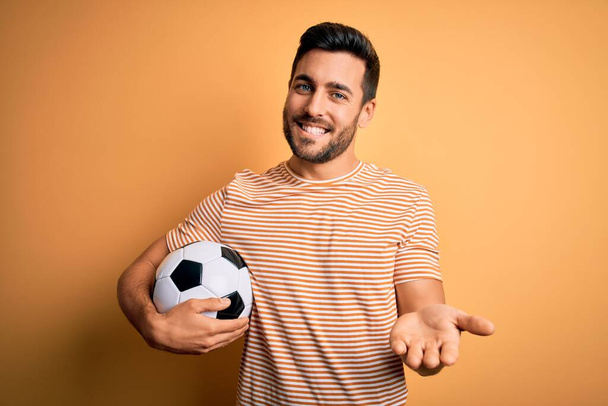 Knappe speler man met baard voetballen houden voetbal over gele achtergrond glimlachend vrolijk aanbieden palm hand geven hulp en acceptatie. - Foto, afbeelding
