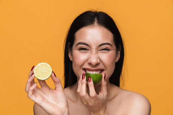 Beauty portrait of caucasian young brunette half-naked woman smiling and eating lemon isolated over yellow background - Foto, immagini