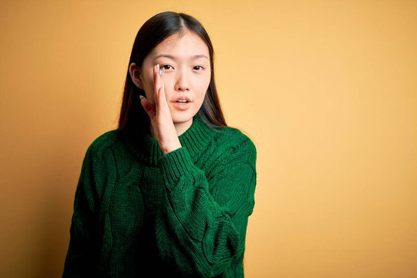 Jonge mooie aziatische vrouw dragen groene winter trui over geel geïsoleerde achtergrond hand op mond vertellen geheim gerucht, fluisteren kwaadaardig gesprek - Foto, afbeelding