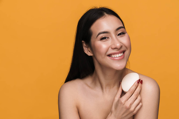 Beauty portrait of young half-naked woman smiling and holding bath soap isolated over yellow background - Foto, Imagen